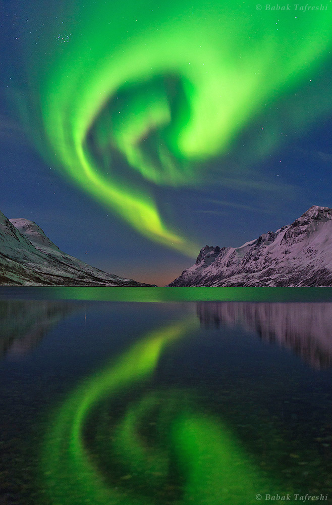 The northern lights or aurora borealis swirls over a fjord in the Norwegian Sea near Tromso, northern Norway. A mix of moonlight, aurora, and a nearby village illuminate the landscape. The Pleiades Star Cluster (the Seven Sisters) appears on the upper left. Credit and copyright: Babak A. Tafreshi.