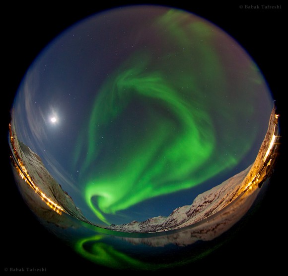 A fisheye view of the northern lights as it swirls over a fjord in the Norwegian Sea near Tromso, northern Norway. A mix of moonlight, aurora, and a nearby village illuminates the the landscape. Credit: Babak A. Tafreshi.