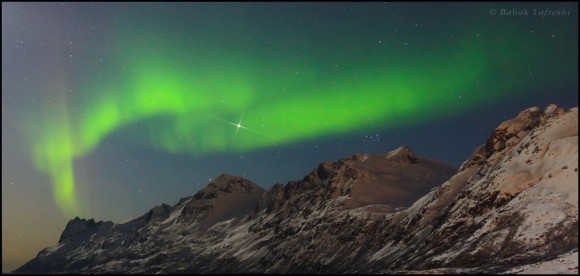 The northern lights or aurora borealis dances over a fjord mountains near Tromso, northern Norway. A mix of moonlight, aurora, and a nearby village illuminate the the landscape. Bright planet Jupiter and the Pleiades Star Cluster (the Seven Sisters) appear over the mountains. Credit and copyright: Babak A. Tafreshi.